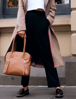 Imagem de um modelo com foco na parte inferior do corpo, usando uma bolsa de cor marrom, usando camiseta curta branca, calça preta e par de calçados pretos