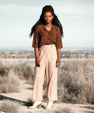 Imagem de uma modelo de etinia negra,de cabelo dread preto, vestindo blusa marrom, calça bege e par de salto alto branco